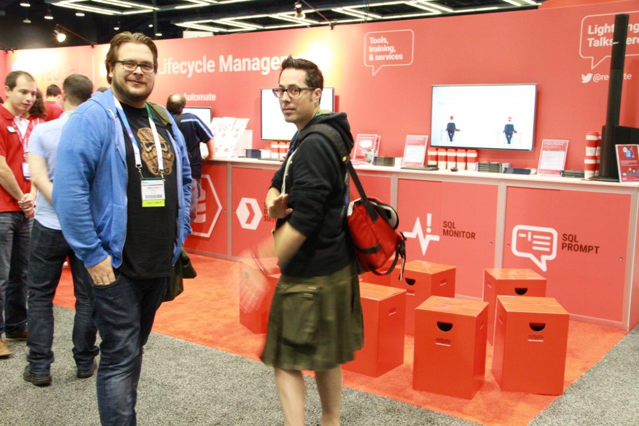 Two male presenting folks are standing in front of the Redgate vendor booth. On the left is a man with light brown hair, wire frame glasses. He is wearing a black t-shirt, blue sweatshirt and blue jeans. Centered in the frame is the post author, turning from the other person towards the camera. They are wearing a green Utilikilt, black sweatshirt and a red messenger bag. They have black hair and black horn rimmed glasses.